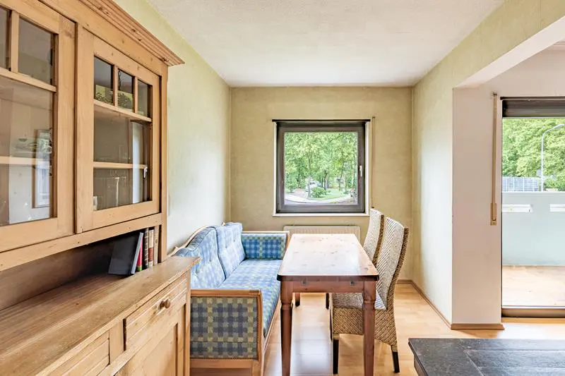 A dining room with a wooden hutch, a blue plaid couch, a wooden table, and two wicker chairs. A window overlooks a green outdoor space.