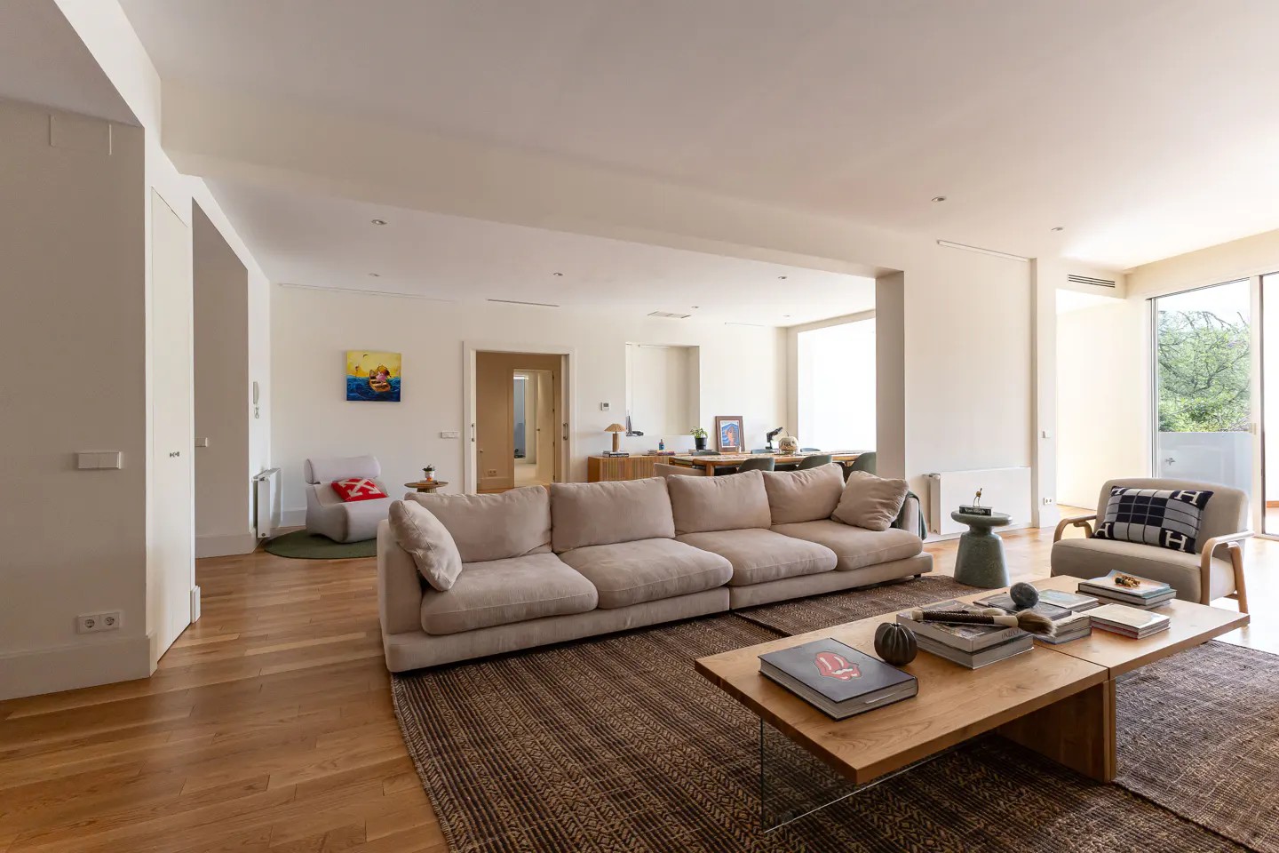 Bright, open living room with a beige sofa, wood floors, and a large coffee table with books. A dining area is visible in the background.
