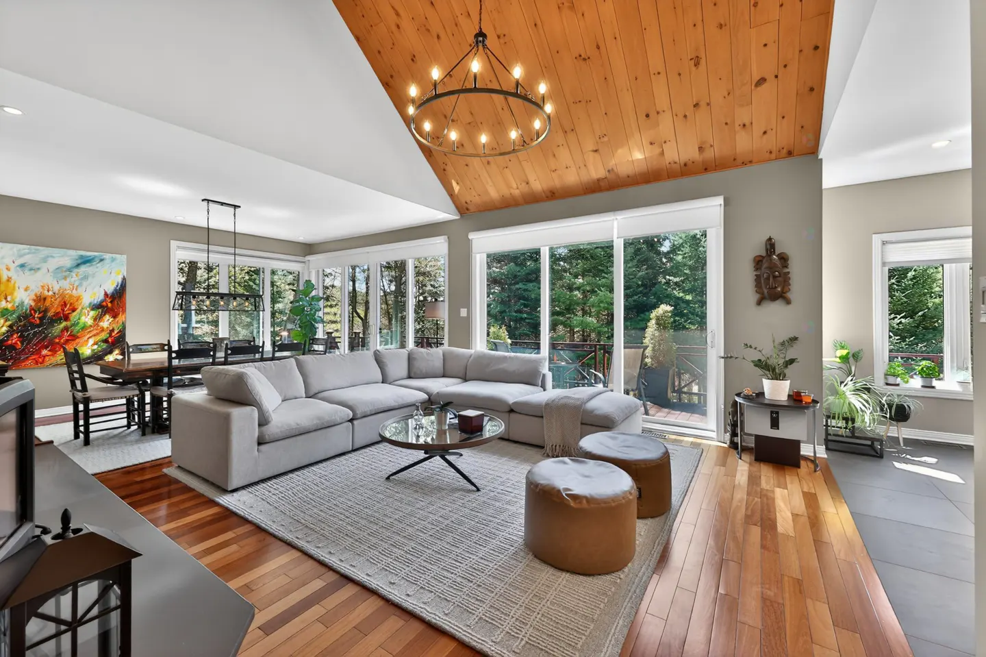 Open-concept living room with wood floors, a gray sectional sofa, and a wood-paneled ceiling with a chandelier. Large windows overlook trees.