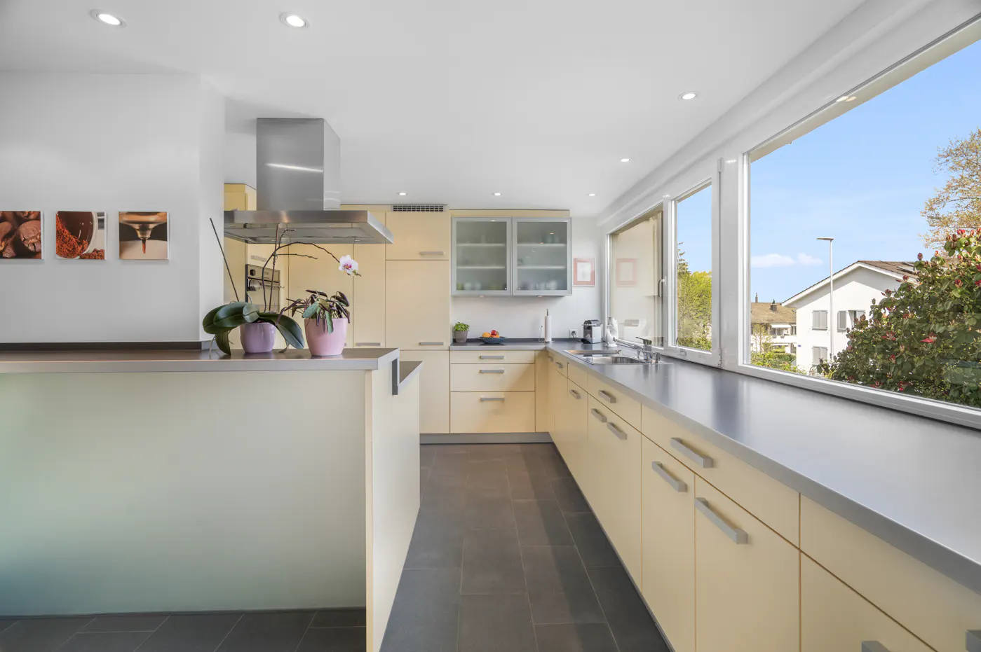 Bright kitchen with light yellow cabinets, stainless steel countertops, and a large window overlooking a residential area.