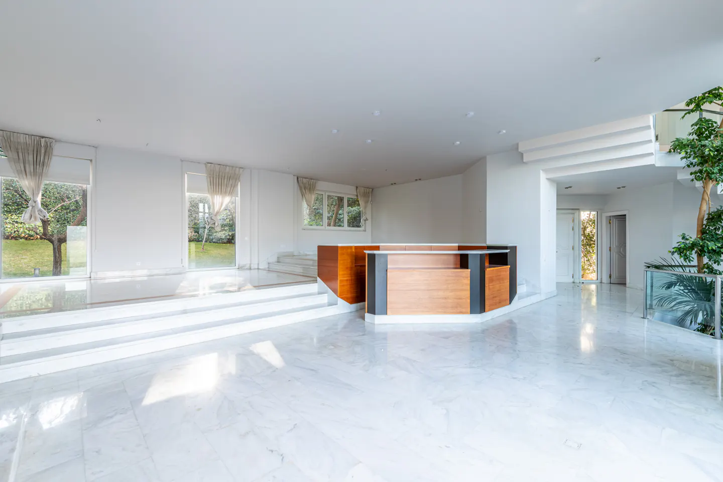 Bright, modern foyer with white marble floors, a wood reception desk, and large windows overlooking a green lawn.