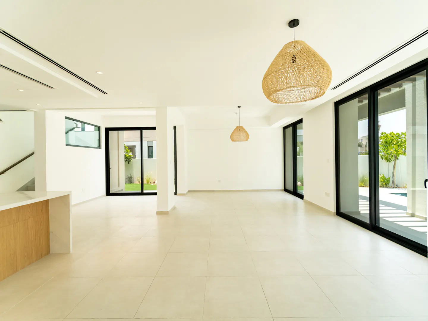Bright, empty living room with white walls, tile floor, and black-framed sliding glass doors to the outside. Wicker pendant lights hang from the ceiling.
