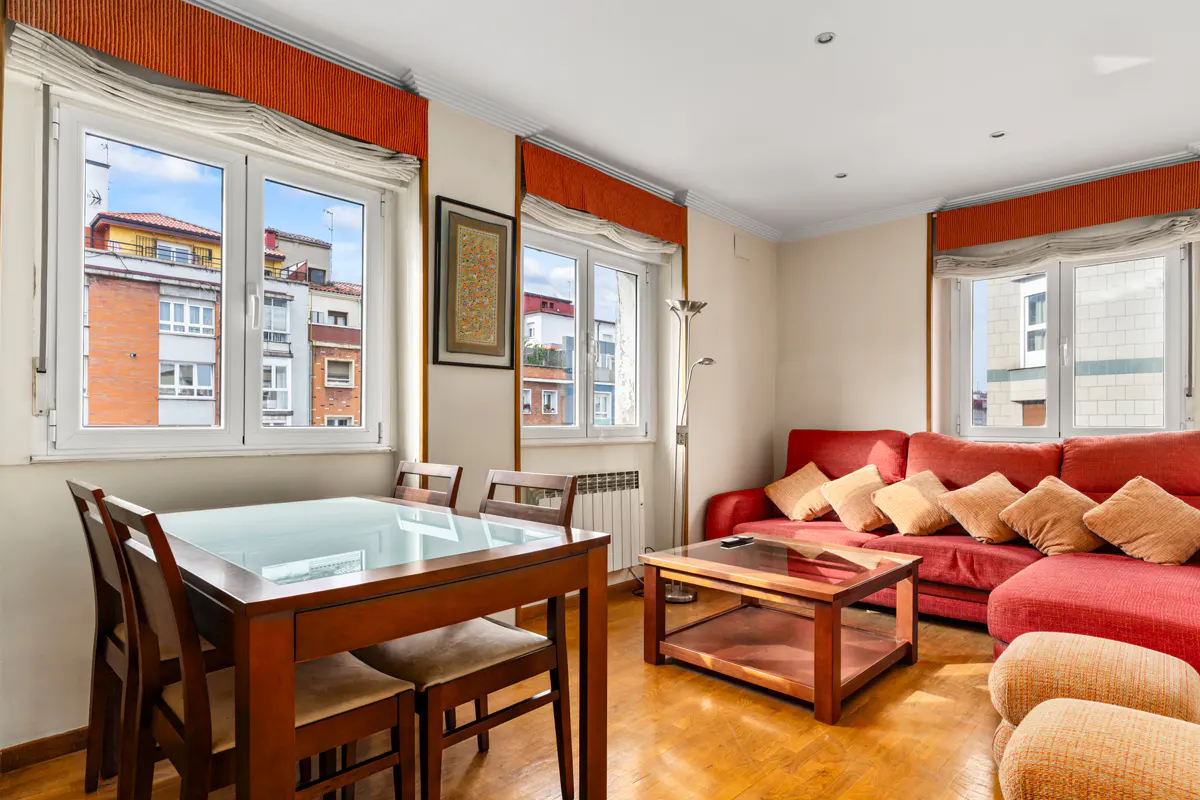A bright living room with a wooden dining table, a red sofa with pillows, and large windows.