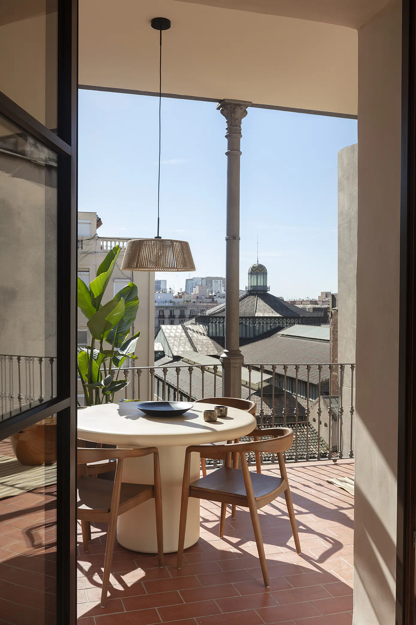 Balcony view with round table, chairs, and woven pendant light. Cityscape visible in the background.