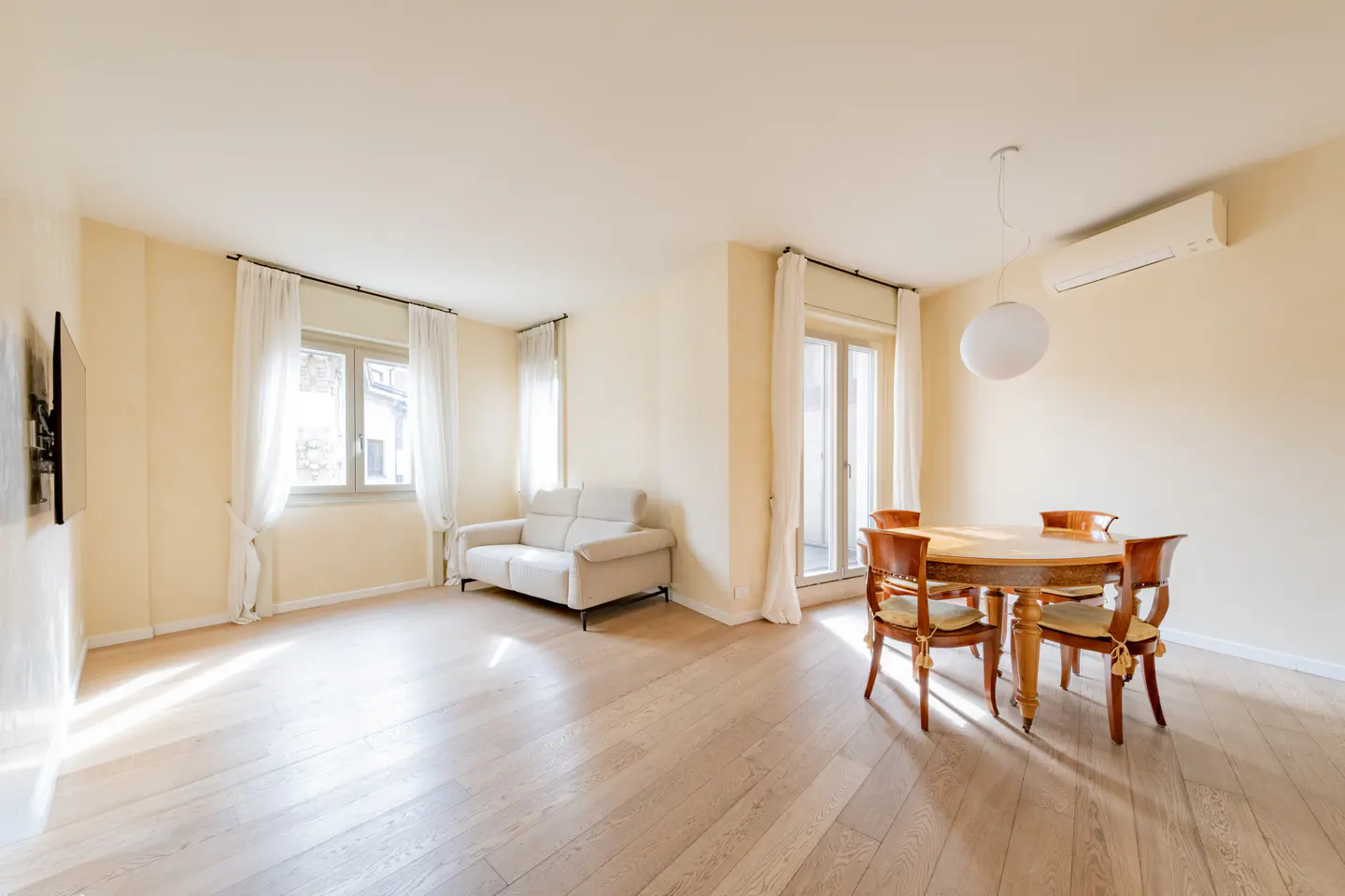 Bright, airy living room with wood floors, a cream sofa, and a round wooden dining table with four chairs. White curtains adorn the windows.