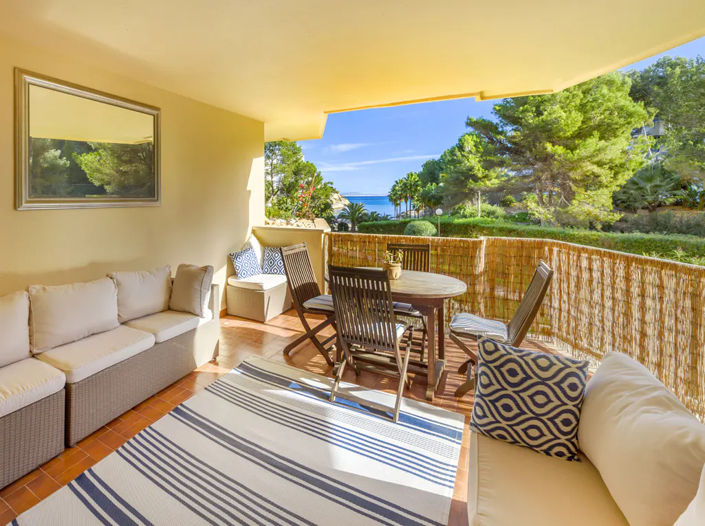 Covered balcony with outdoor sofa, dining set, and striped rug, overlooking a sunny coastal landscape with trees and sea.