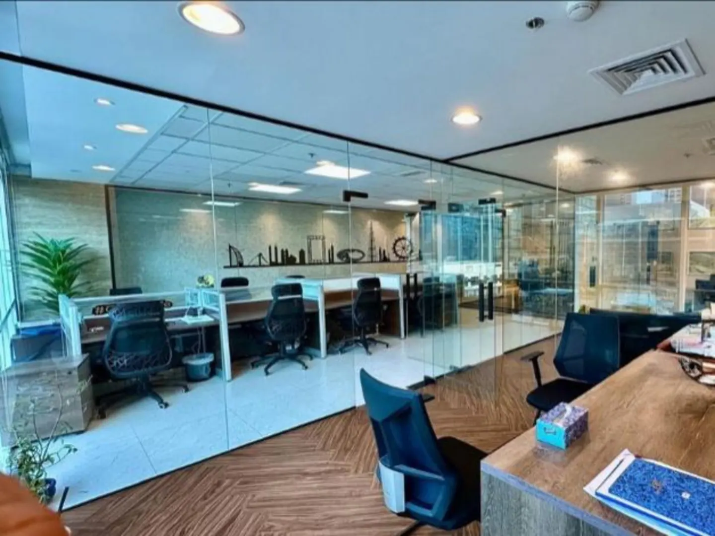 Bright office space with glass walls, desks, and black chairs. A city skyline graphic adorns the wall. Wood-patterned flooring adds warmth.