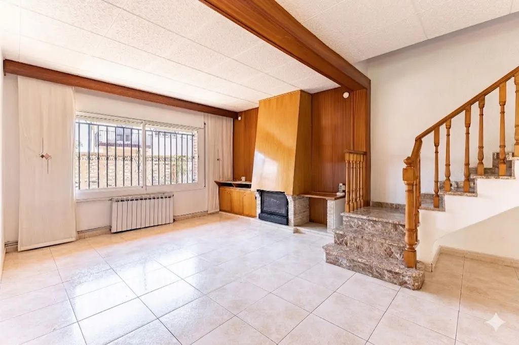 A living room with tile floors, a wood-paneled fireplace, and a staircase with marble steps. A window with white curtains is on the left.