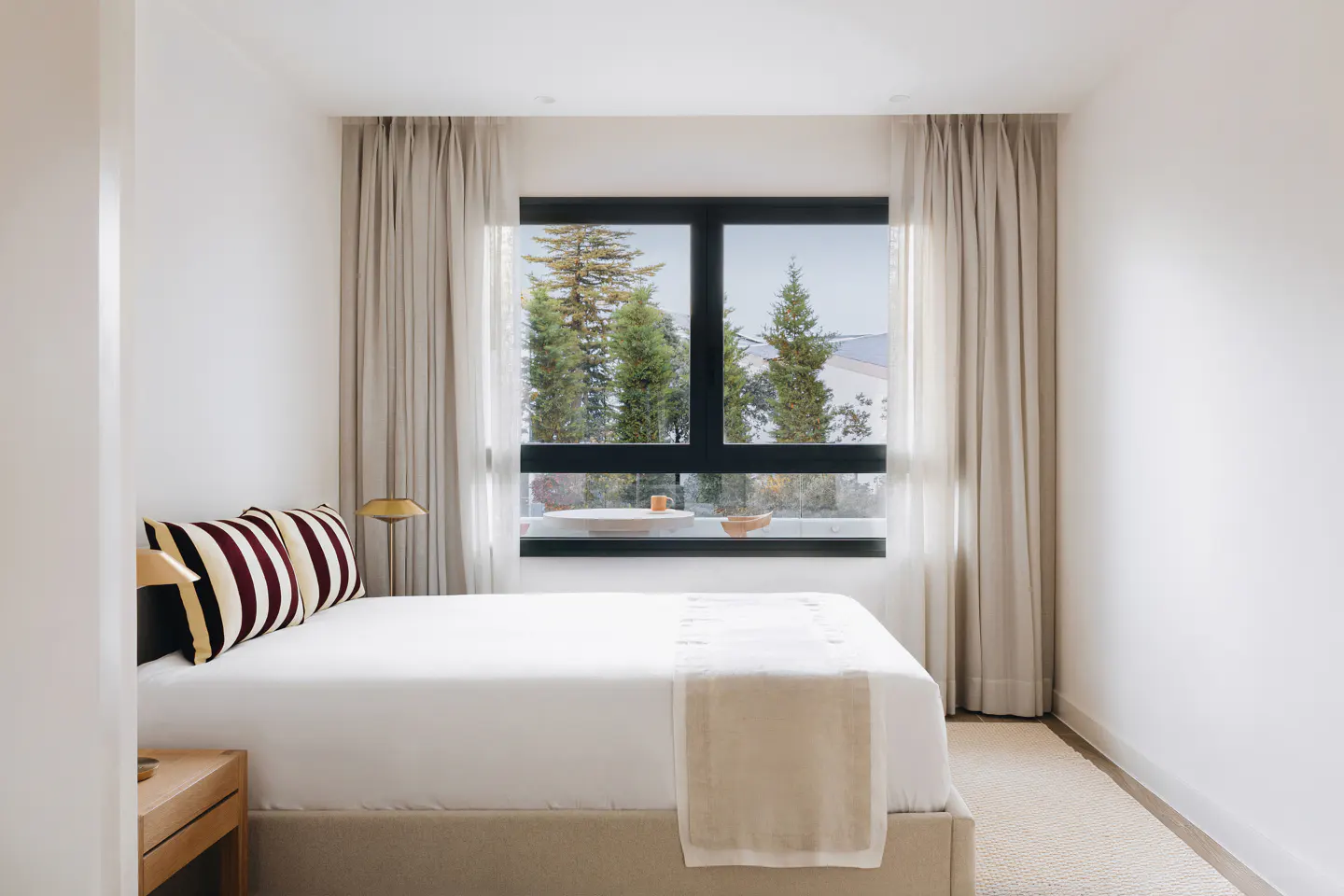 Bright bedroom with white walls, a bed with striped pillows, and a black-framed window showing trees.