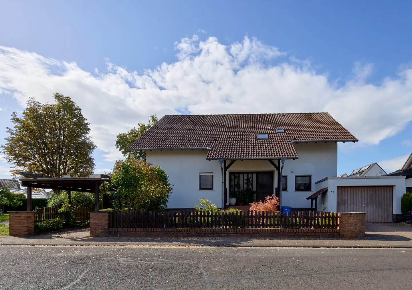 Two-story white house with a brown roof, a wooden fence, and a blue sky with white clouds.