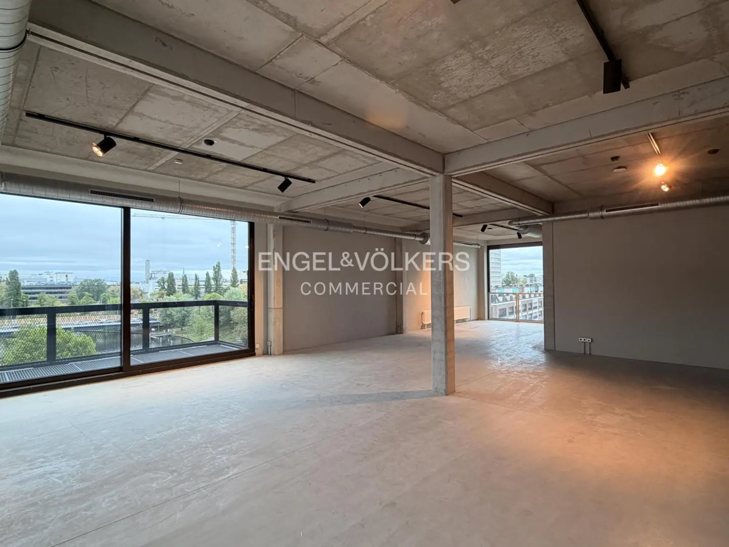 An empty commercial space with concrete floors, ceiling, and support beams. Large windows offer a city view. Spotlights illuminate the room.