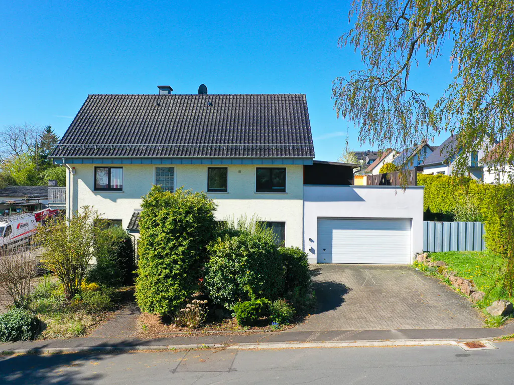 Two-story house with a gray roof, white walls, and a white garage door. Green bushes in front and a blue sky above.