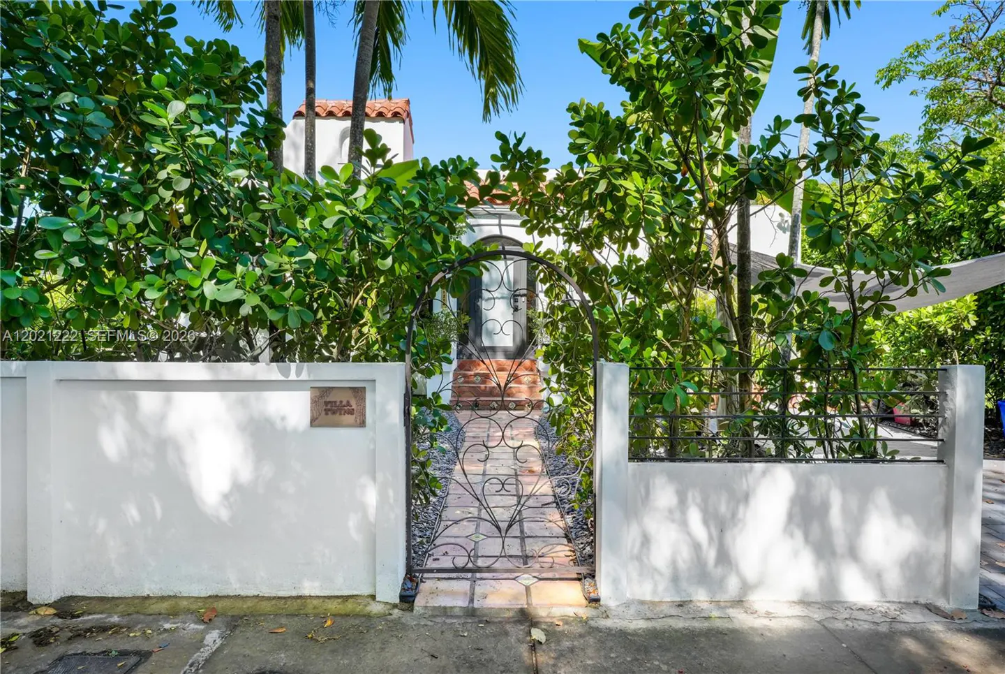 Gated entrance to a white house with a red tile roof, surrounded by lush green trees and plants. "Villa Twins" sign on the left wall.