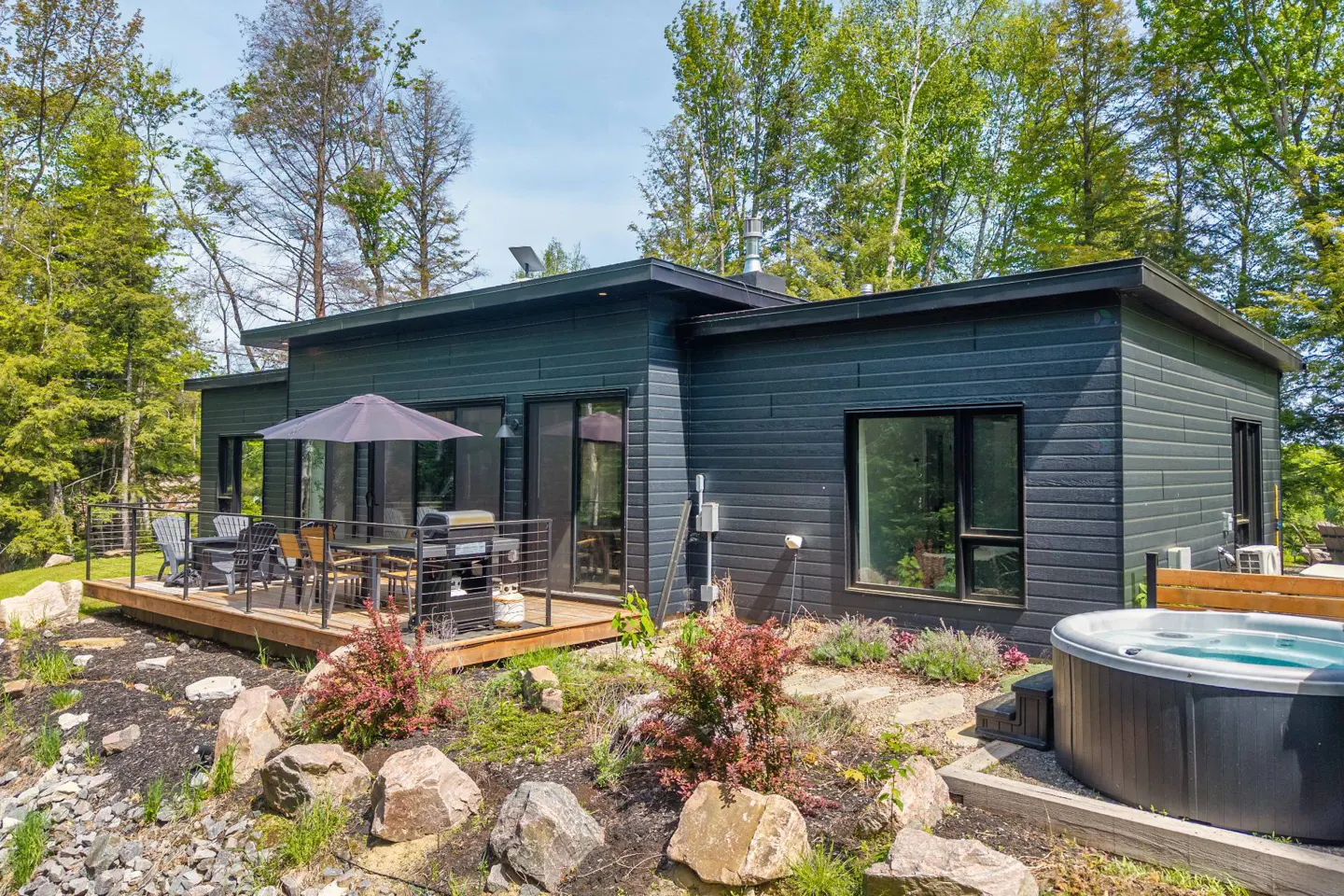 Modern dark-sided home with a wooden deck, patio furniture, umbrella, and a hot tub in a lush, wooded backyard.