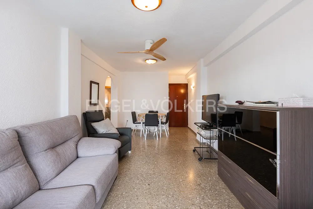 Living room with a grey sofa, black armchair, dining table with four chairs, and a TV on a stand with terrazzo flooring.