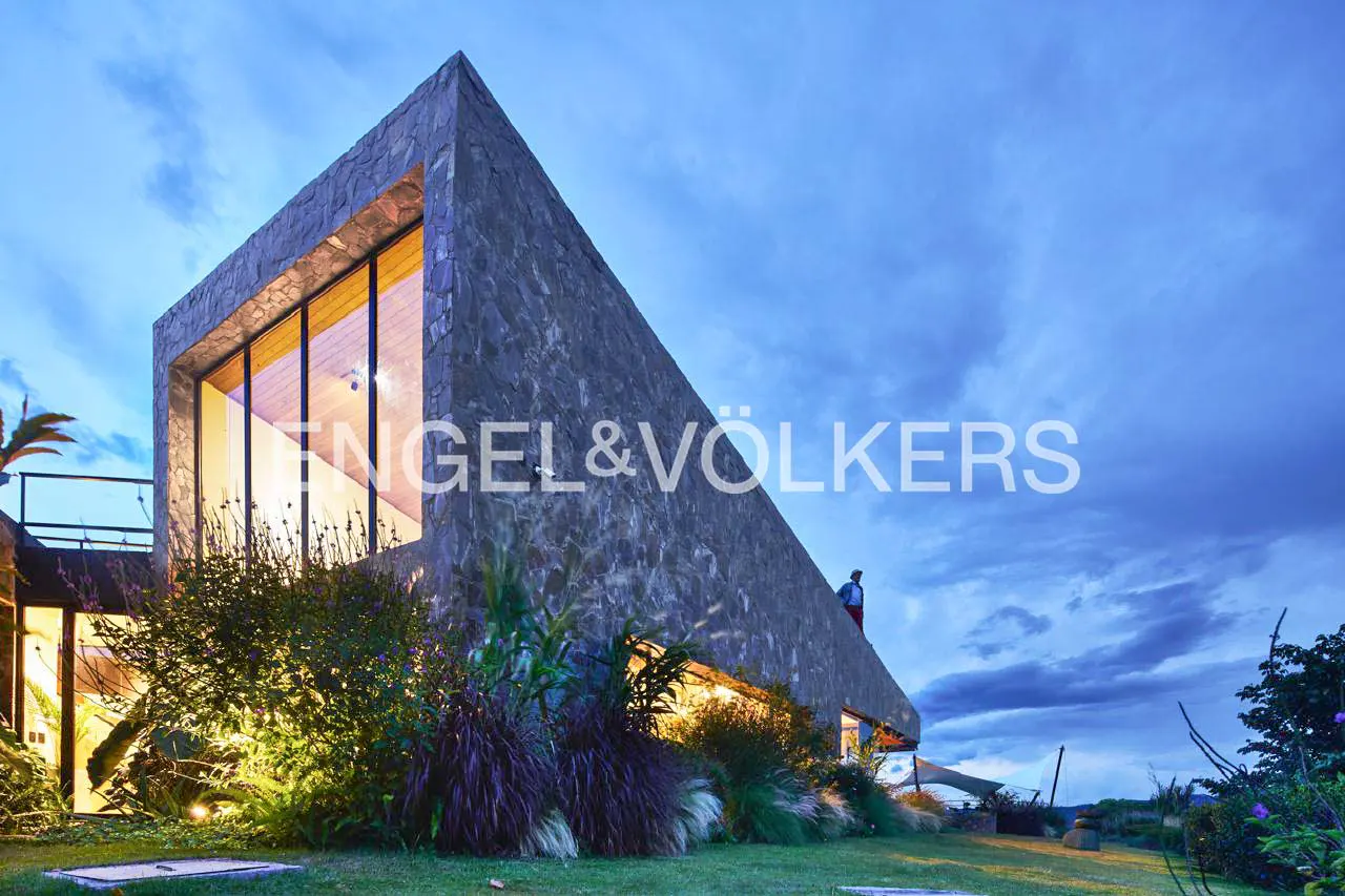 Modern stone-clad house with large glass windows at twilight, surrounded by lush landscaping under a blue cloudy sky.
