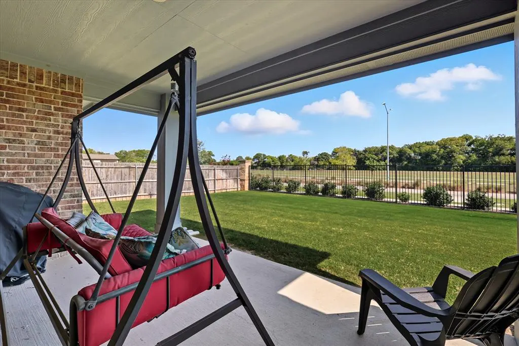 Covered patio with a red swing seat and pillows, a black chair, and a view of a green lawn and trees.