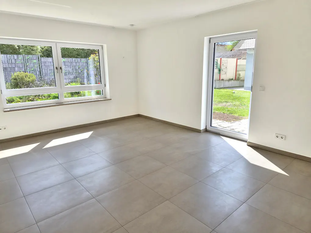 Bright, empty room with gray tile floor, white walls, window overlooking green yard, and open glass door to the outside.