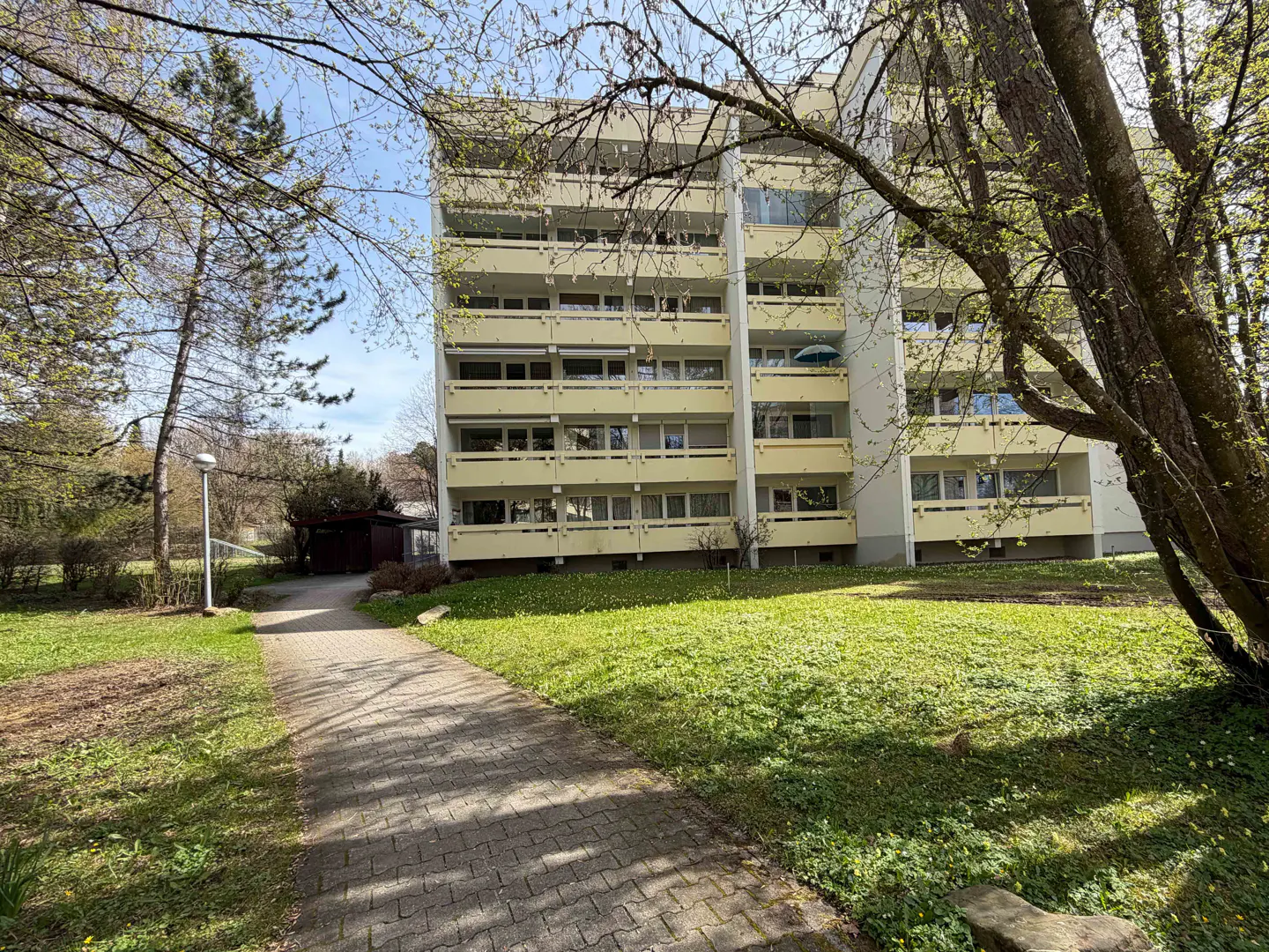 Exterior view of a light yellow apartment building with balconies, a brick walkway, and green grass.