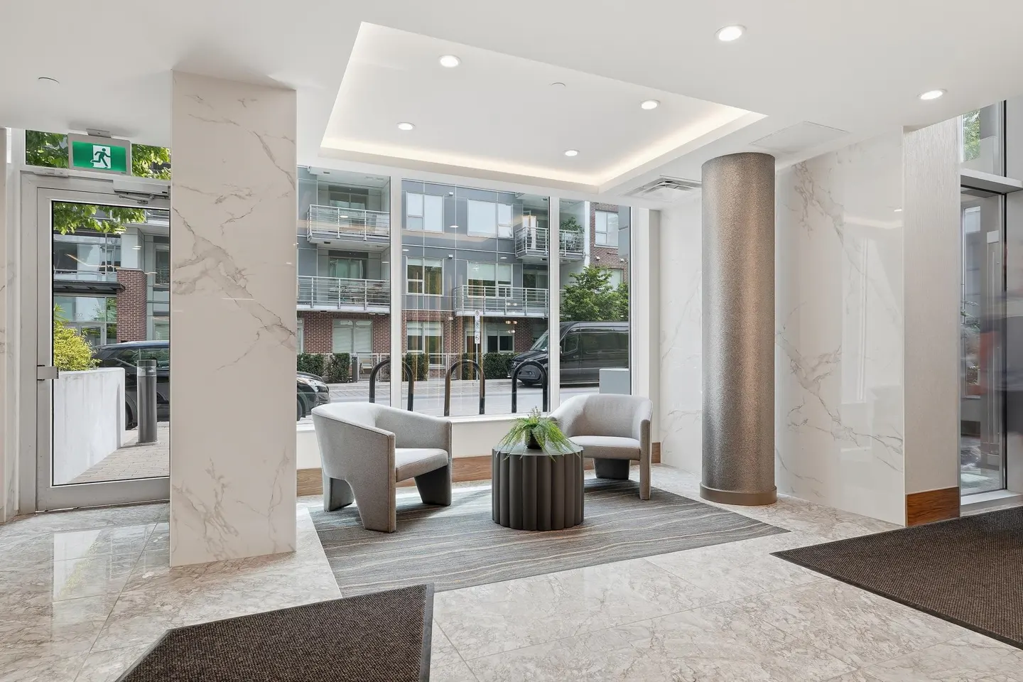 Lobby area with two gray chairs, a round table with a plant, and a large window with a view of a building.