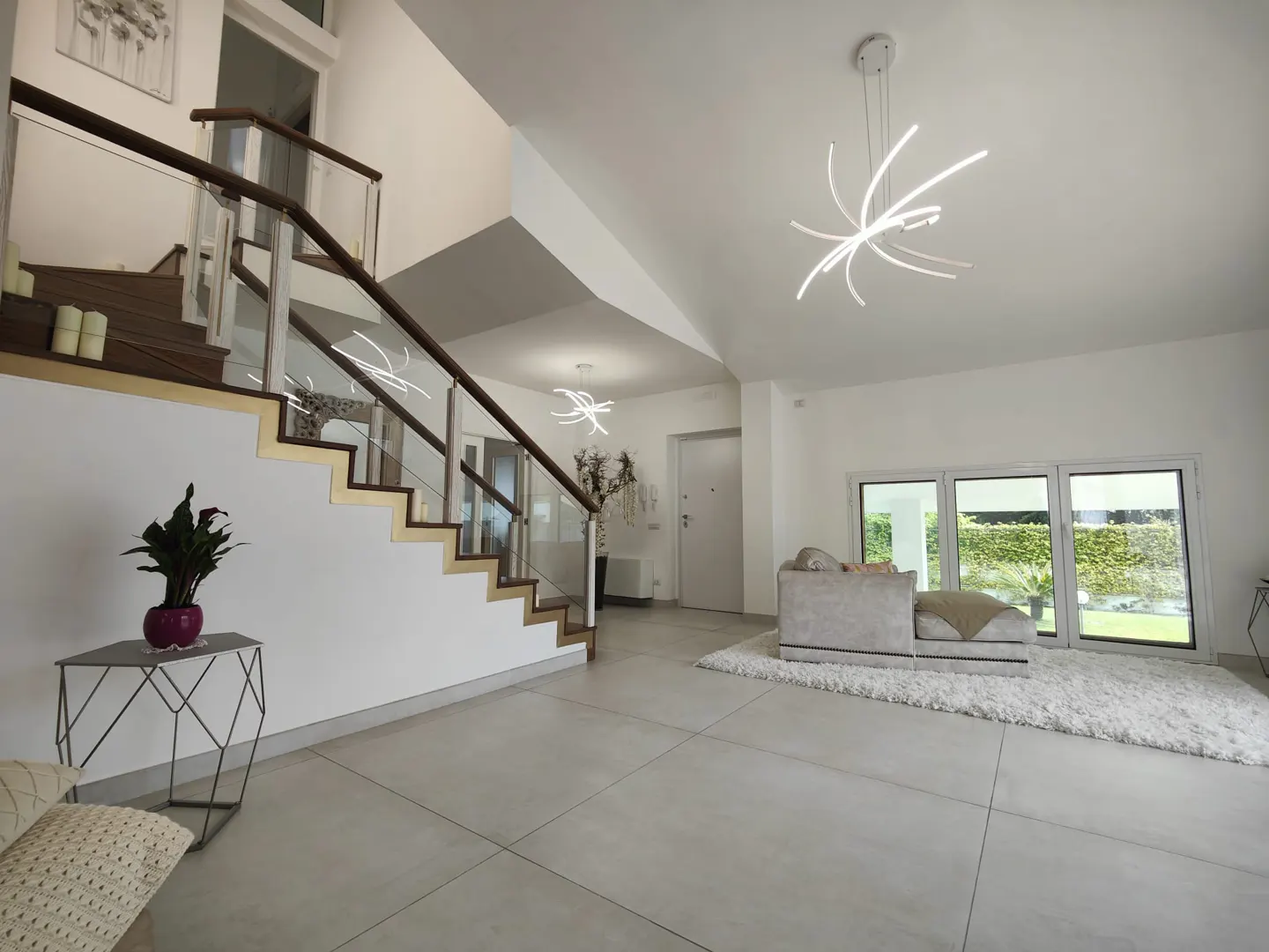 Bright, modern foyer with a staircase, glass railings, and a unique light fixture. A white shag rug sits near a window overlooking a green yard.