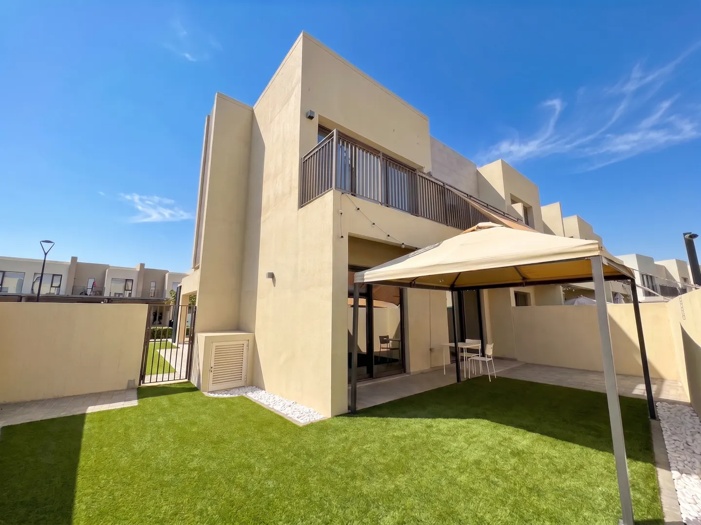 Beige two-story home with a balcony, green lawn, and a beige gazebo in the backyard under a clear blue sky.
