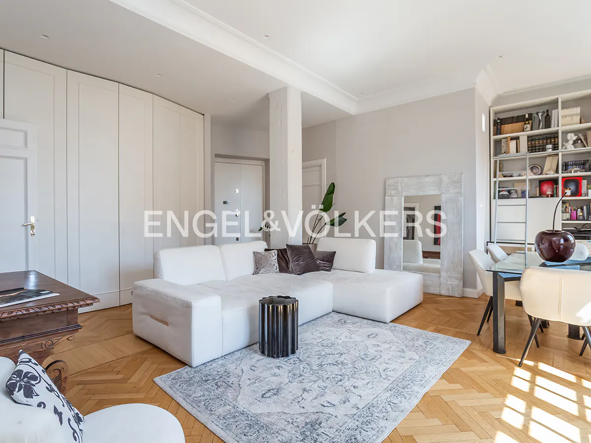 Bright living room with white sectional sofa, patterned rug, and herringbone wood floors. Dining table and bookshelf in background.