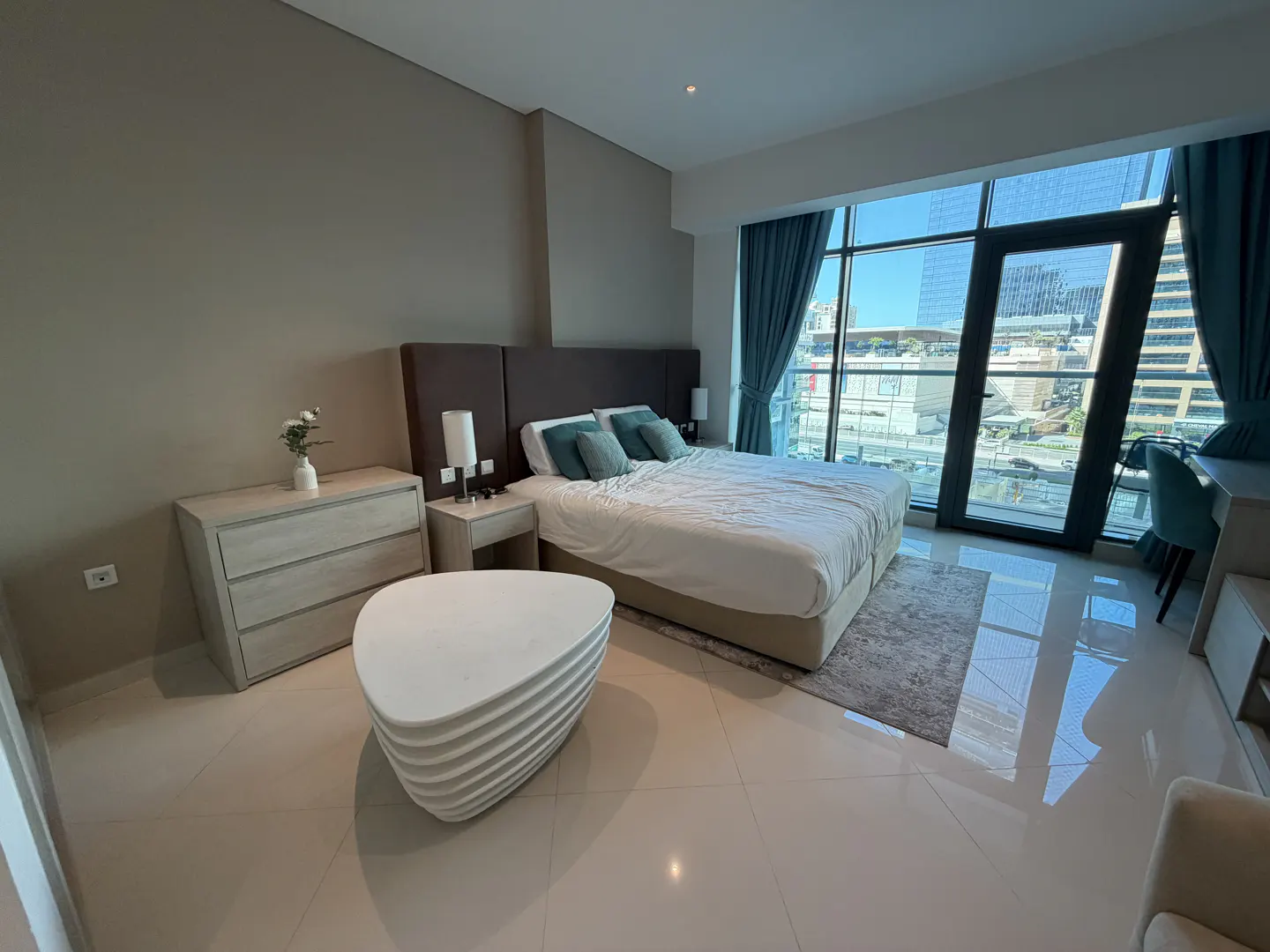 Modern bedroom with a white bed, unique tiered coffee table, light wood dresser, and floor-to-ceiling city view windows.