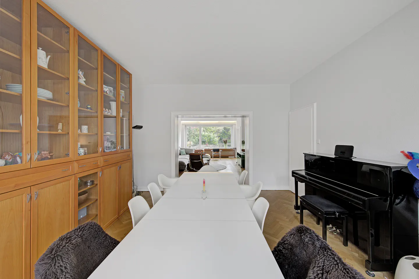 Bright dining room with a long white table, white chairs, and a black piano. A wooden cabinet displays dishware. A living room is visible beyond.