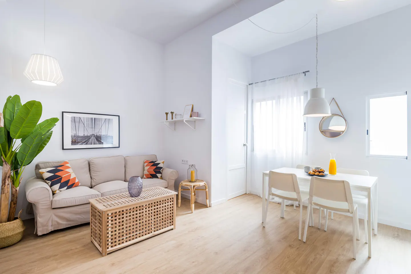Bright, modern living and dining area with beige sofa, wood table, and white dining set. A large green plant adds a pop of color.