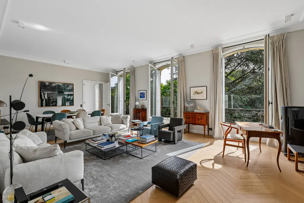 Bright, spacious living room with hardwood floors, grey sofas, and large windows overlooking lush greenery. Neutral color palette.