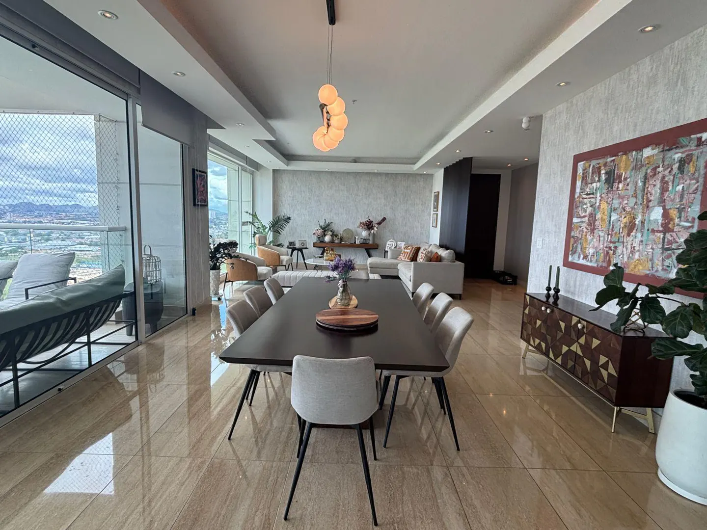 A wide shot of a modern dining and living room with a large table, chairs, and a balcony with a city view.