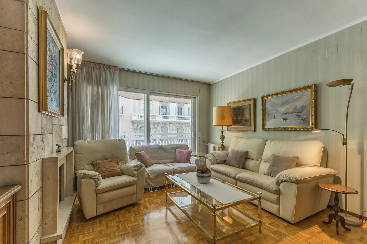 Living room with parquet floor, cream sofas, glass coffee table, and paintings on the pale green walls. Balcony view in background.