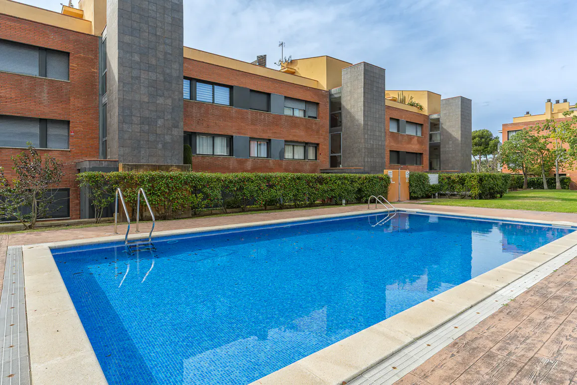 Outdoor pool with blue tile, surrounded by green hedges and brick apartment buildings under a cloudy sky.