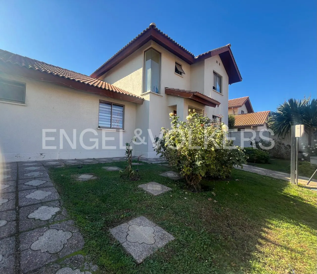 Two-story beige house with a brown roof, green lawn, and stone walkway under a blue sky. Engel & Volkers logo on the house.