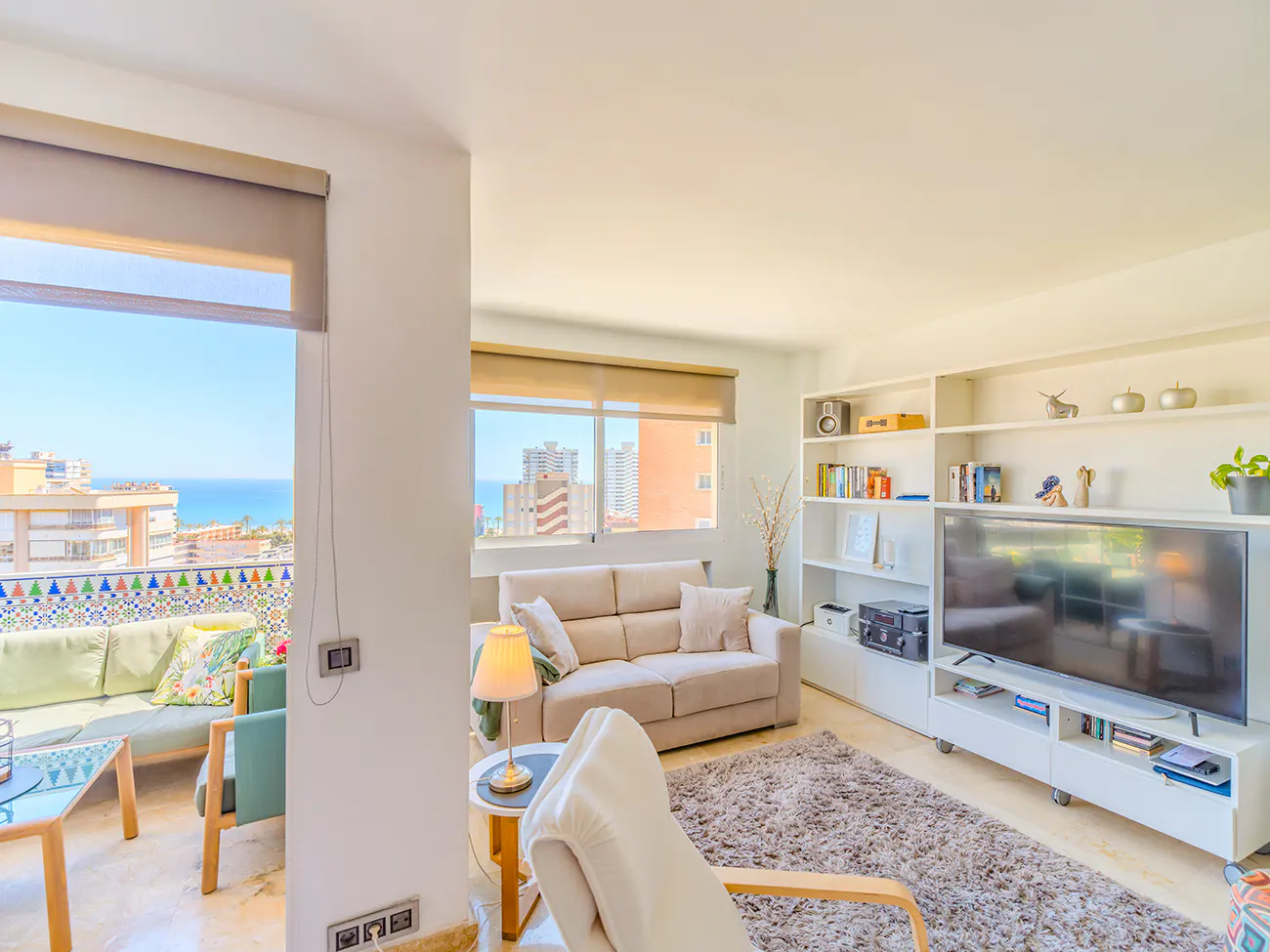 Bright living room with beige sofa, white shelves, and TV. Balcony with ocean view visible through window.
