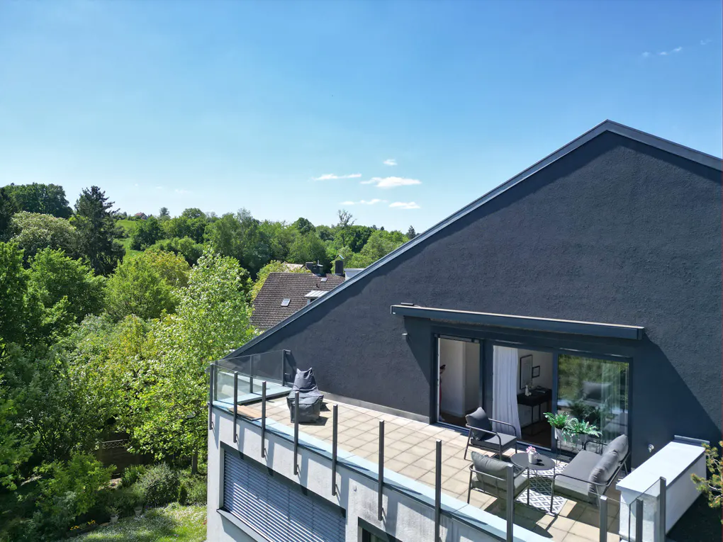 Balcony with gray furniture, glass railing, and sliding glass doors on a dark gray building, overlooking green trees under a blue sky.