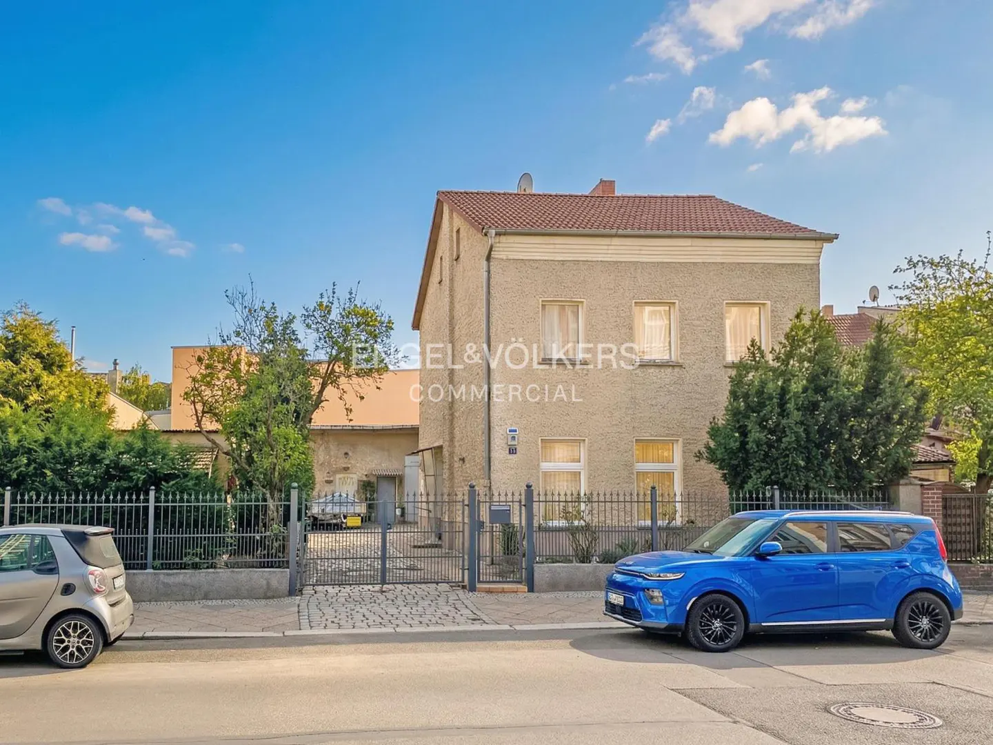 Two-story beige building with a red tile roof, a blue car parked in front, and a gray car parked on the street.