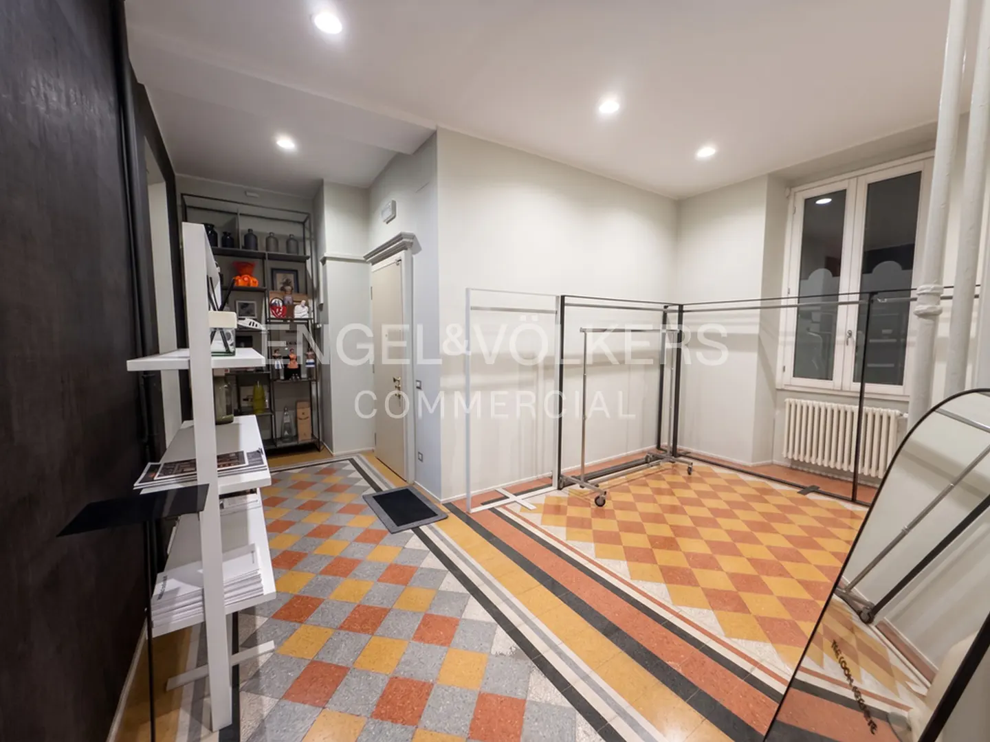 Bright commercial space with checkered tile floors, metal clothing racks, white shelving, and a window with a radiator.