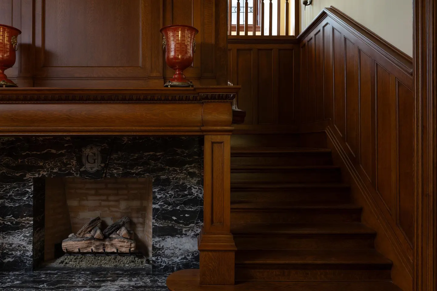 Interior view of a wood-paneled foyer with a marble fireplace and a staircase. Two red vases sit on the mantelpiece.