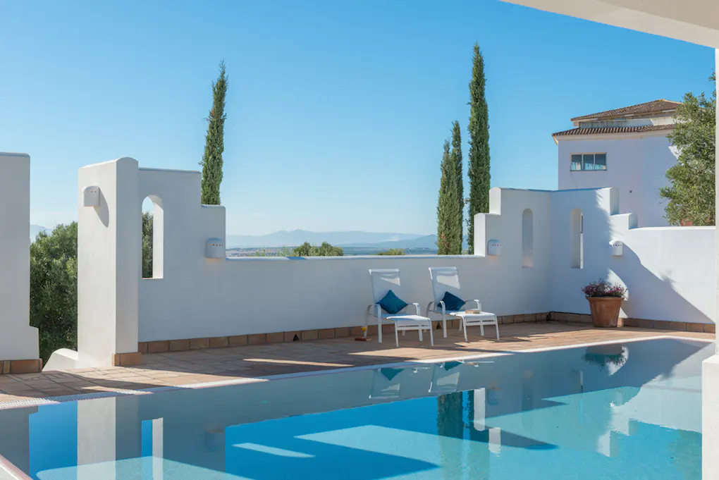 A bright, sunny view of a blue swimming pool with two white lounge chairs and a white building in the background.