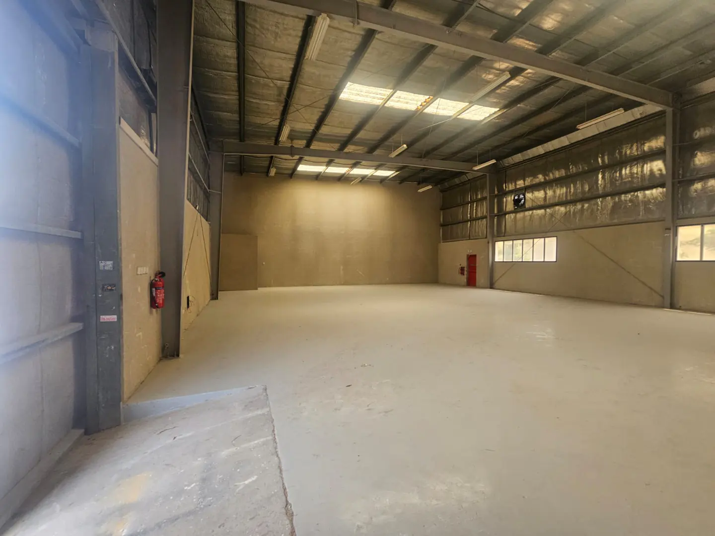 Empty warehouse interior with gray concrete floor, metal ceiling, and a red fire extinguisher on the wall.