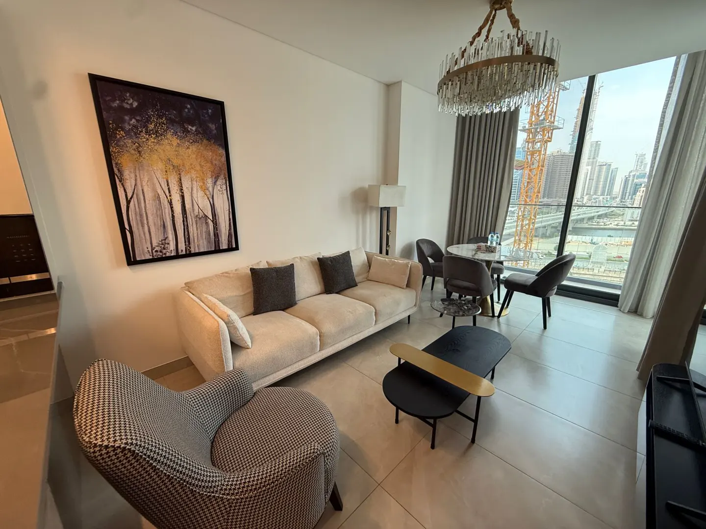 Living room with beige sofa, patterned chair, and black coffee table. A framed tree art hangs on the wall. City view through large windows.