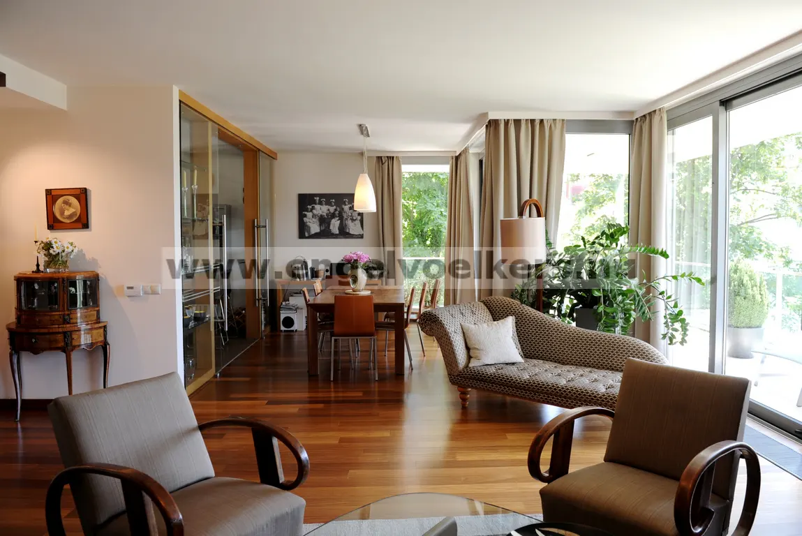 A bright living room with hardwood floors, two armchairs, a chaise lounge, and a dining table. Large windows let in natural light.