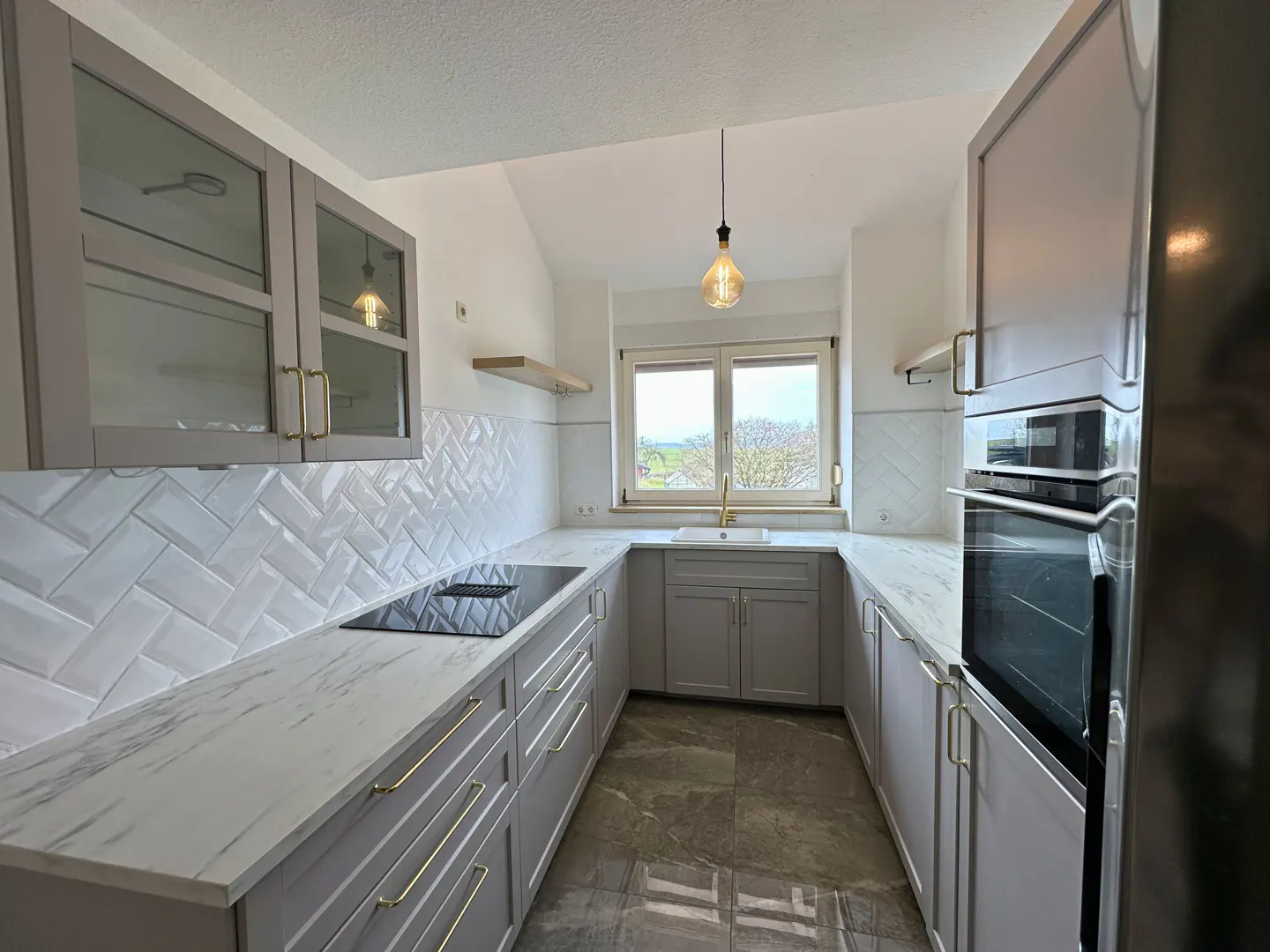 Bright kitchen with gray cabinets, marble countertops, and herringbone tile backsplash. A window overlooks a green landscape.