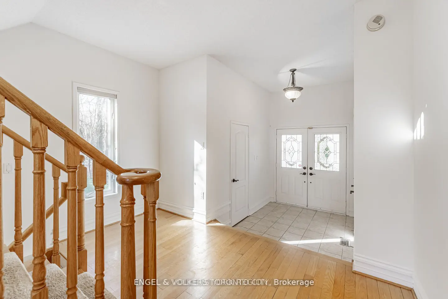 Bright foyer with white walls, hardwood floors, and a staircase with a wooden banister. Double doors with decorative glass lead to the outside.