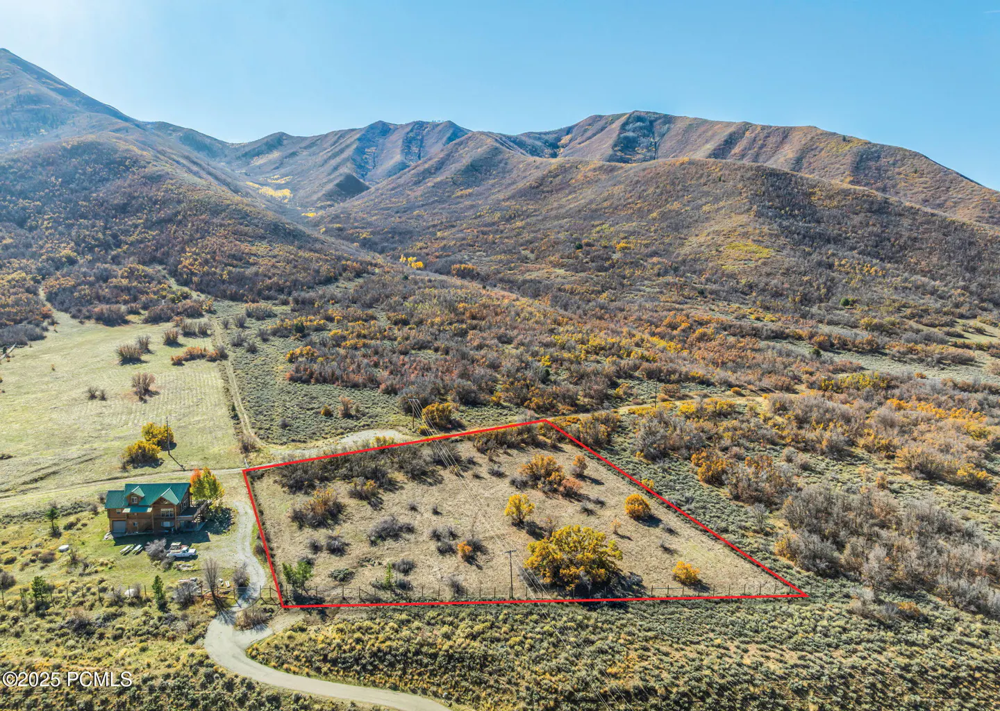 Aerial view of a vacant lot outlined in red, with a house, mountains, and blue sky in the background.