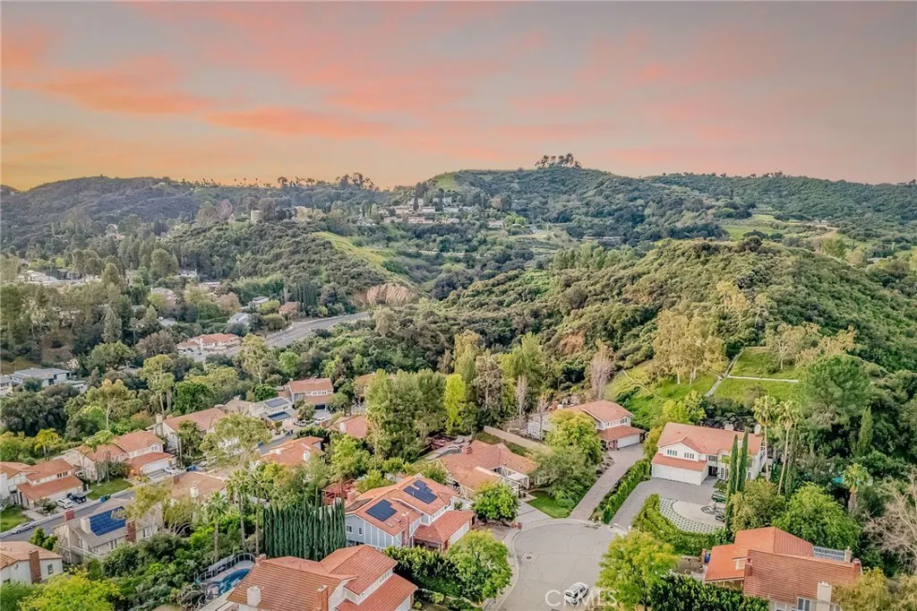 Aerial view of a residential neighborhood with houses with red tile roofs, surrounded by green hills and trees under a pink and orange sky.