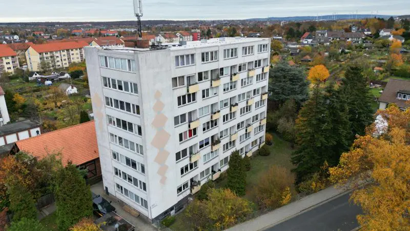 Aerial view of a tall, white apartment building with many windows and small balconies, surrounded by trees and houses.