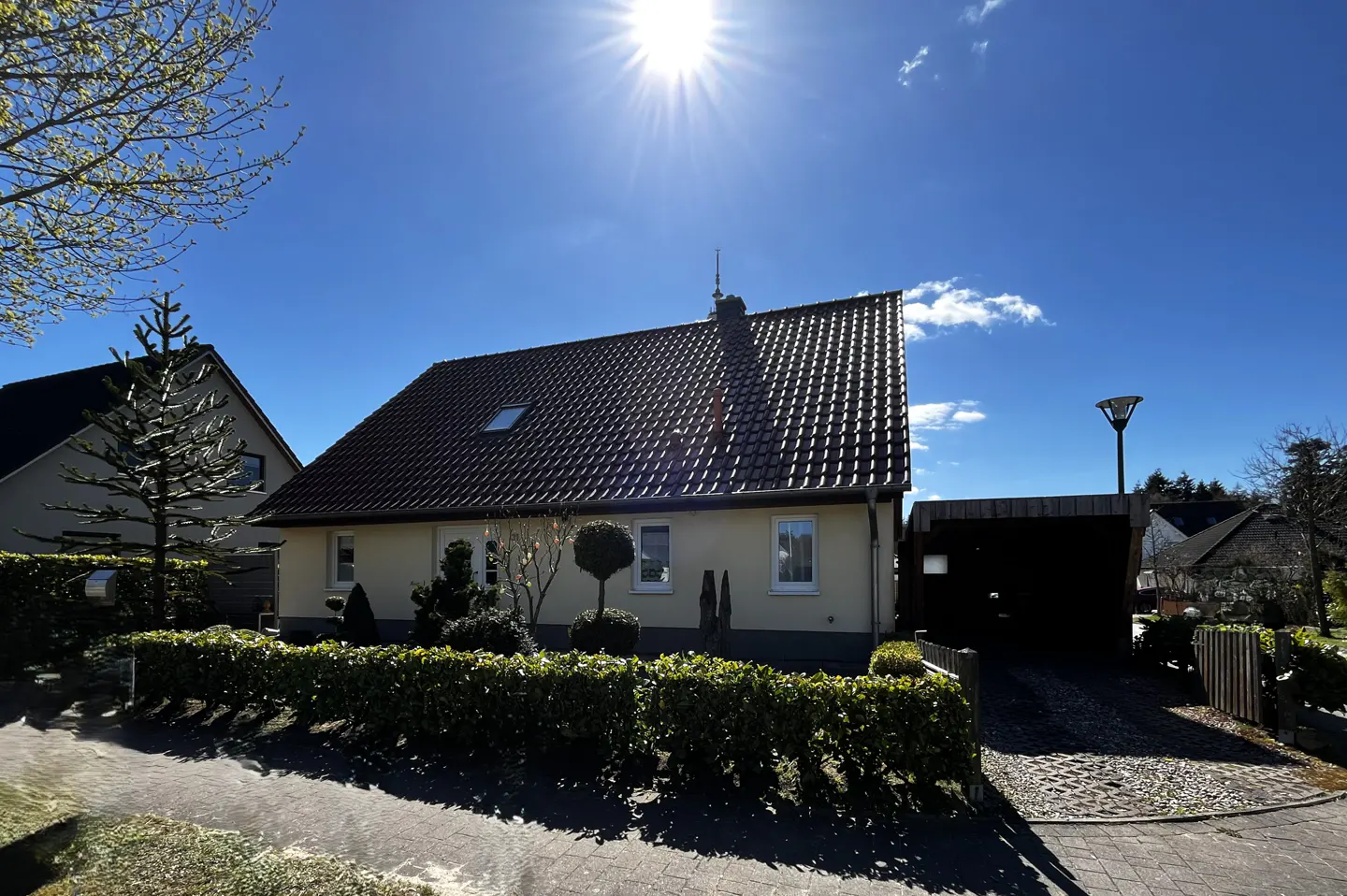 Exterior view of a one-story beige house with a dark brown roof under a bright blue sky and sun. A green hedge lines the front.